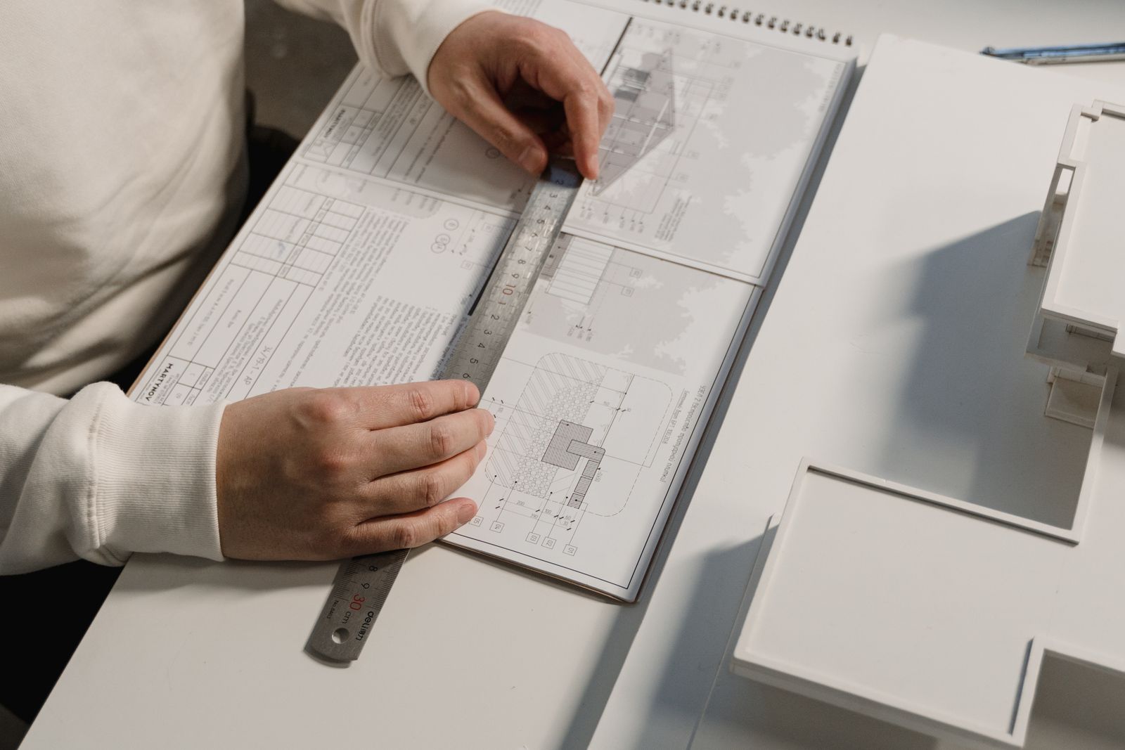 Mains posées sur un jeu de plans techniques, règle métallique et maquette blanche sur un plan de travail
